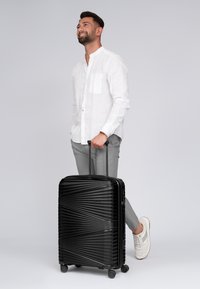 Hombre con camisa blanca y pantalones grises sosteniendo el mango de una maleta rodante negra con diseño texturizado, de pie contra un fondo liso.