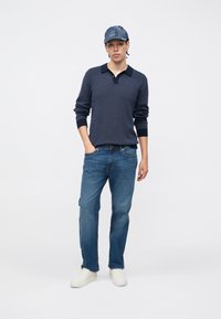 Marineblaues Polohemd mit langen Ärmeln und kontrastierendem Kragen, blaue Jeans aus Denim und weiße Slip-On-Schuhe, ergänzt durch eine blaue gemusterte Mütze.