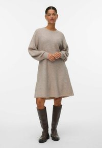 Frau steht in einem beigefarbenen gestrickten Langarmkleid und dunkelbraunen kniehohen Stiefeln vor einem schlichten weißen Hintergrund.