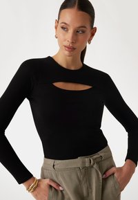 Top lungo a maniche nere a coste con un'apertura sul colletto. Il modello lo indossa infilato in pantaloni khaki a vita alta con una cintura.