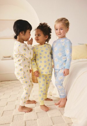 Kolme pientä lasta kukallisissa pyjamissa seisoo paljain jaloin kuvioidulla matolla pehmeästi valaistussa makuuhuoneessa.