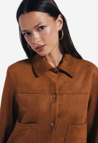 Brun mockaläderjacka med krage, framknäppning med knappar och två framfickor. Slät yta med subtila sömnadsdetaljer.