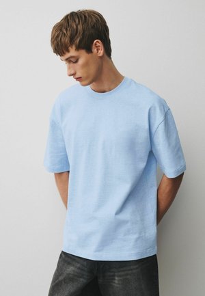 Junger Mann mit hellbraunen Haaren, trägt ein hellblaues übergroßes kurzärmeliges T-Shirt und dunkle Jeans, blickt nach unten und hat die Hände hinter dem Rücken.