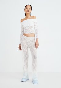 Bijela čipkasta majica s kratkim rukavima i off-shoulder dizajnom, u kombinaciji s prozirnim bijelim čipkastim hlačama. Oboje imaju teksturirani uzorak. Plave platforme upotpunjuju odjevnu kombinaciju.