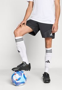 Μαύρα ποδοσφαιρικά παπούτσια πάνω σε μια μπάλα με μπλε και λευκά σχέδια. Ο παίκτης φοράει λευκές κάλτσες με μαύρες ρίγες και μαύρο σορτς με υφασματική υφή.