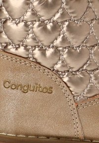 Primer plano de tela acolchada beige con patrón de corazones y logotipo "Conguitos" bordado en dorado sobre material de textura suave.