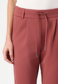 Roestkleurige broek met een trekkoord in de taille, zijzakken en zachte, gestructureerde stof. Plooien voegen visueel belang toe aan het ontwerp.