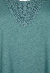 Tissu sarcelle avec un motif triangulaire en dentelle au crochet présentant des motifs floraux et circulaires près de l'encolure.