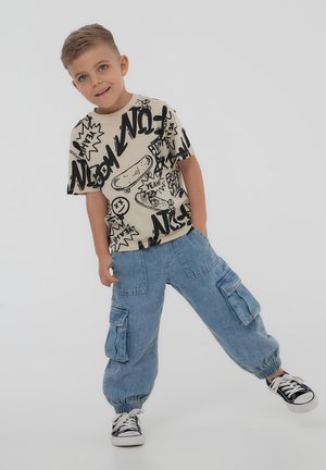 Maglietta beige con motivi e illustrazioni grafiche nere, abbinata a pantaloni cargo in denim azzurro chiaro con polsini elastici e molteplici tasche.