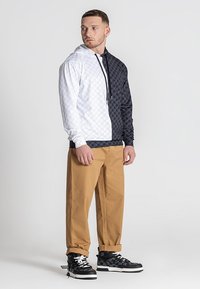 Džemper s kapuljačom u dizajnu s blokovima boja; bijela s jedne strane s suptilnim uzorkom, crna s druge strane. Uparen s svijetlosmešim hlačama i crnim cipelama.