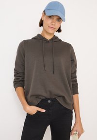 Felpa marrone con cappuccio e coulisse, vestibilità comoda e polsini a coste, abbinata a jeans neri. Un berretto azzurro chiaro completa il look informale.