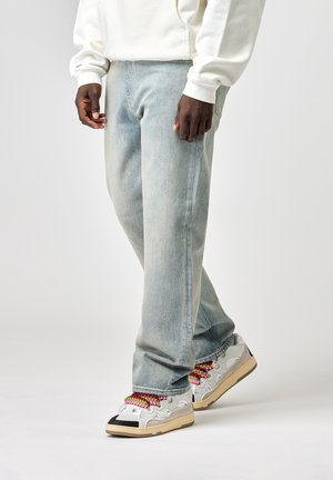 Personne portant un jean bleu clair, des baskets blanches et noires avec des lacets colorés, et un sweat à capuche blanc, debout devant un fond uni.