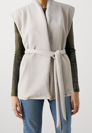 Gestrikt vest in lichtgrijs gestreepte stof met een ceintuur in de taille, voorzien van een kraagloos ontwerp en open mouwen. Draagt over een groene top.