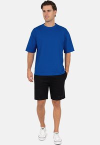 T-shirt en coton bleu à manches courtes associé à un short noir, avec un logo discret. Porté avec des baskets blanches, sur un fond uni.