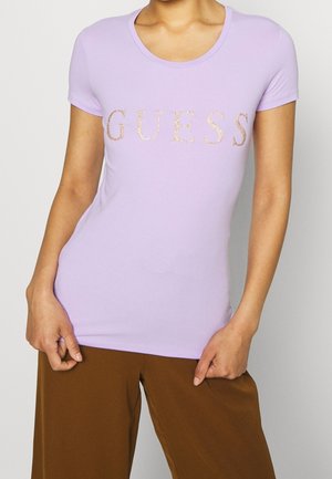 Kobieta ubrana w dopasowaną lawendową koszulkę z krótkim rękawem z błyszczącym napisem „GUESS”, w połączeniu z luźnymi brązowymi spodniami, pokazana od szyi do bioder.