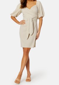 Beige rutmönstrad omslagsklänning med puffärmar och en frontknut, knä-lång, i kombination med nude slide-sandaletter. Slät tygtextur.