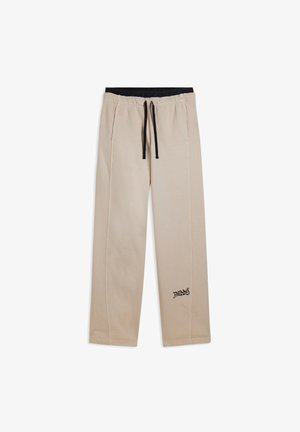Pantalones casuales beige con cordón negro, cinturilla elástica, bolsillos laterales y pequeño logo negro "Freddy" cerca del tobillo izquierdo.