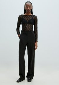 Schwarzer Spitzen-Bodysuit mit langen Ärmeln, V-Ausschnitt und sichtbaren Unterwäschedetails, kombiniert mit hochtaillierten schwarzen Hosen.