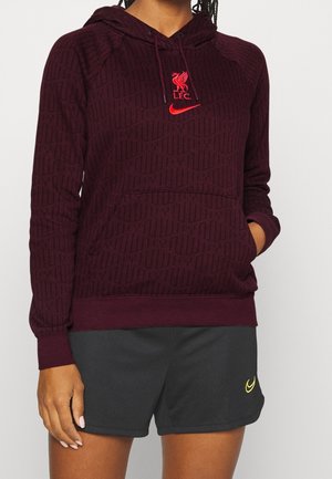 Person iført en mørkerød Liverpool FC-hoodie med rød logo og sorte Nike-shorts med en lille gul swoosh.