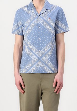 Mężczyzna noszący niebieską koszulę z krótkim rękawem zapinaną na guziki z białym paisley bandana printem i oliwkowo-zielone spodnie.