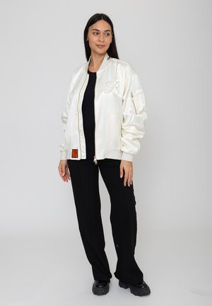 Femme portant une veste bomber blanche brillante sur un t-shirt noir, un pantalon noir et des chaussures noires épaisses, debout devant un fond clair uni.