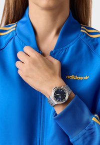 Kék poliészter dzseki sárga csíkokkal és Adidas logóval. Ezüst óra kék számlappal és fém hálós pánttal a csuklón.