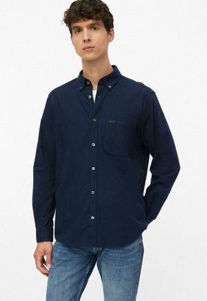Junger Mann, der ein dunkelblaues Hemd mit Brusttasche und hellblaue Jeans trägt, steht vor einem schlichten weißen Hintergrund.