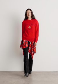Rød strikket sweater med broderet design, parret med en sort og rød ternet skjorte bundet om livet og sorte bukser, båret med sko.