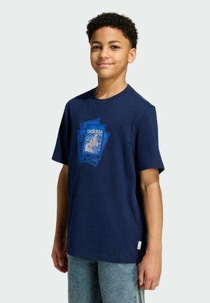 Smiling boy with short curly hair wearing a navy blue Adidas T-shirt with graphic cards design and light blue jeans, standing against a plain background.
