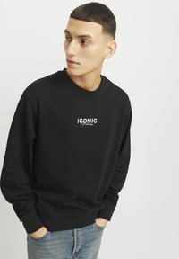Sudadera negra con cuello redondo y mangas largas, con el logo blanco "ICONIC". Combinada con jeans de color azul claro, de tejido sutilmente texturizado.
