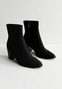 Botas negras hasta el tobillo hechas de un tejido suave, con punta afilada, tacón cilíndrico y cremallera lateral para facilitar su uso.