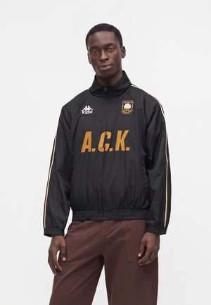 Hombre con chaqueta deportiva negra con cremallera con estampado amarillo A.C.K., logo de Kappa y emblema, combinado con pantalones marrones, sobre fondo blanco liso.