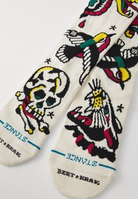 Cotton socks featuring colorful designs of a skull, bird, and other motifs on a cream background, with black and teal accents.