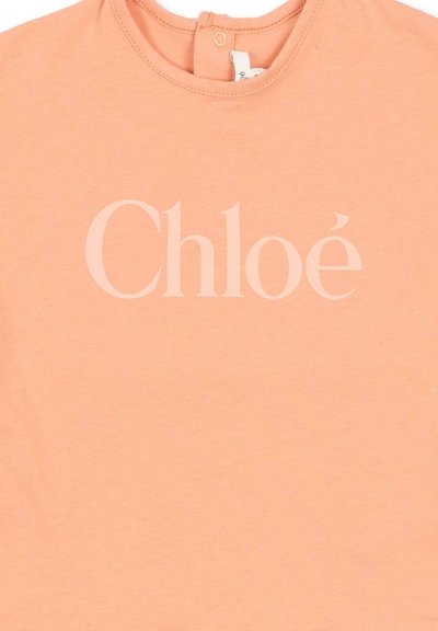 Brzoskwiniowa koszula z okrągłym dekoltem i zapięciem na guzik z tyłu, z nazwą marki "Chloé" nadrukowaną dużymi, jasnoróżowymi literami.