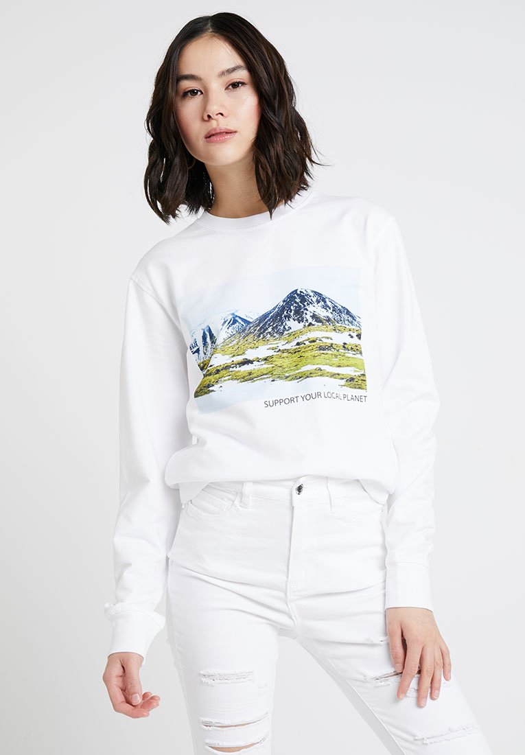 Biała bluza z grafiką górskiego krajobrazu i napisem "WSPIERAJ SWOJĄ LOKALNĄ PLANETĘ". W połączeniu z poszarpanymi białymi dżinsami.