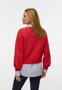 Jersey corto rojo con mangas abullonadas, tejido texturizado, dobladillo acanalado. Lleva puesto sobre una camisa de collar azul claro y combinado con jeans oscuros.