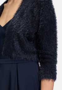 Veste cropped en fausse fourrure bleu marine avec des manches trois-quarts, offrant une finition douce et pelucheuse ainsi qu'une silhouette ajustée.