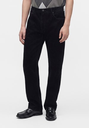 Person trägt schwarze, gerade geschnittene Hose und schwarze Leder-Loafer, die Hände entspannt an den Seiten, vor einem schlichten Hintergrund.
