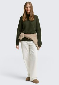 Dunkelgrüner, gerippter Pullover mit langen Ärmeln, beiges Kleidungsstück, das in der Taille gebunden ist, kombiniert mit weißen weiten Hosen und braunen Slippern.