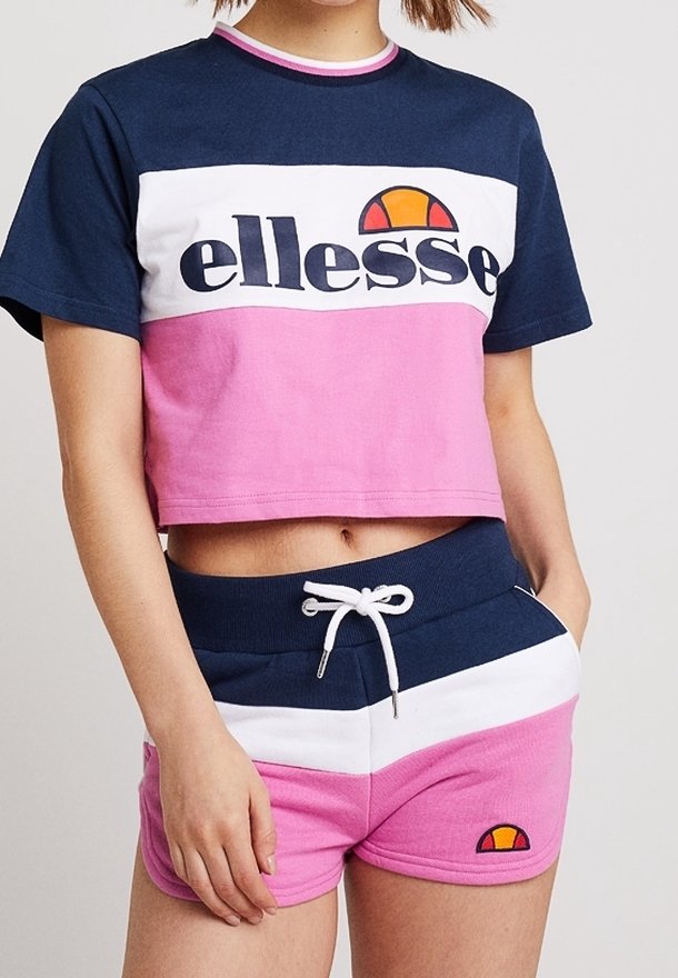 Femme portant un crop top ellesse bleu marine, blanc et rose ainsi qu'un short assorti avec une taille à cordon et des écussons avec logo.