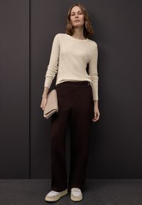 Maglione crema a coste a maniche lunghe, pantaloni marrone scuro a gamba larga e clutch beige intrecciata. Le scarpe da ginnastica grigie con dettagli crema completano l'outfit.