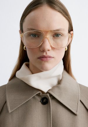 Jongere met lang lichtbruin haar draagt grote, doorzichtige bril, gouden oorringen, beige jas en een witte top met hoge hals tegen een witte achtergrond.