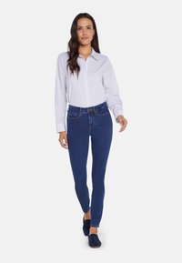 Camisa blanca de botones con finas rayas azules, combinada con jeans ajustados de color azul oscuro y zapatos slip-on en navy, que cuentan con un collar clásico y un diseño ajustado.