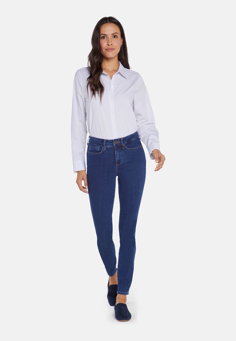 Camisa blanca de botones con finas rayas azules, combinada con jeans ajustados de color azul oscuro y zapatos slip-on en navy, que cuentan con un collar clásico y un diseño ajustado.