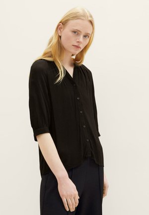 Zwarte blouse met knoopsluiting en korte pofmouwen, textuurstof en een V-hals. G gedragen met donkere broeken, die een ontspannen pasvorm toont.