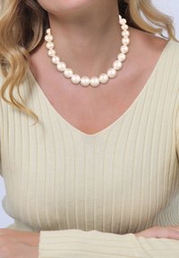Collier en perles avec de grosses perles crémeuses et brillantes, disposées en un motif symétrique, porté sur un haut beige clair texturé.