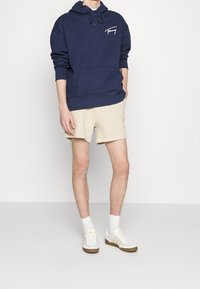 Marinblå huvtröja med framficka och vit logotyp, tillsammans med beiga shorts. Vita sneakers med beiga detaljer och vita strumpor.