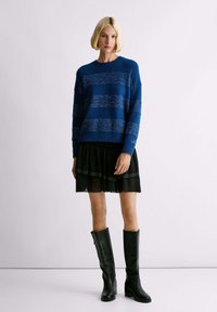 Suéter a rayas azules con acentos metálicos, superpuesto a una falda negra de volantes, combinado con botas altas de cuero negro. Textura suave y corte relajado.