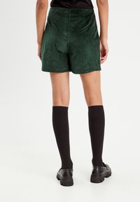 Green corduroy shorts with a high waist and textured fabric, paired with black knee-high socks and black shoes, viewed from the back.