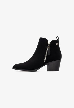 Botín de ante negro hasta el tobillo con punta afilada, detalle de cremallera lateral y tacón apilado. Presenta una pequeña lengüeta en la parte posterior.